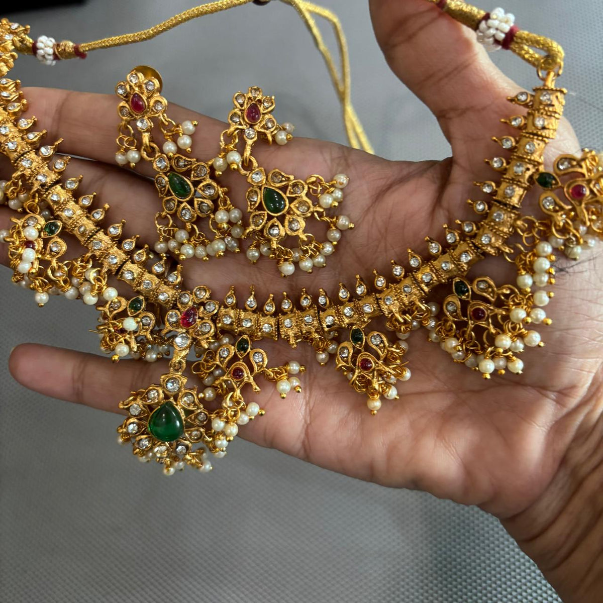Handheld close up of traditional Gold-tone diamond style necklace set with green centre stone, ruby accents and pearls cluster