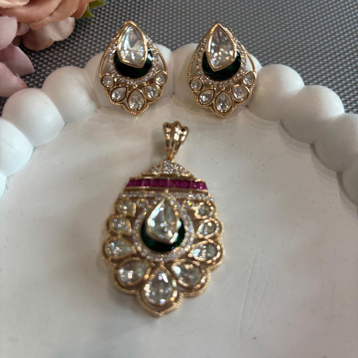 Gold-toned traditional Indian pendant set featuring a teardrop-shaped pendant and matching stud earrings, embellished with clear stones, a dark green enamel , and a row of red stones, displayed on a white scalloped tray.