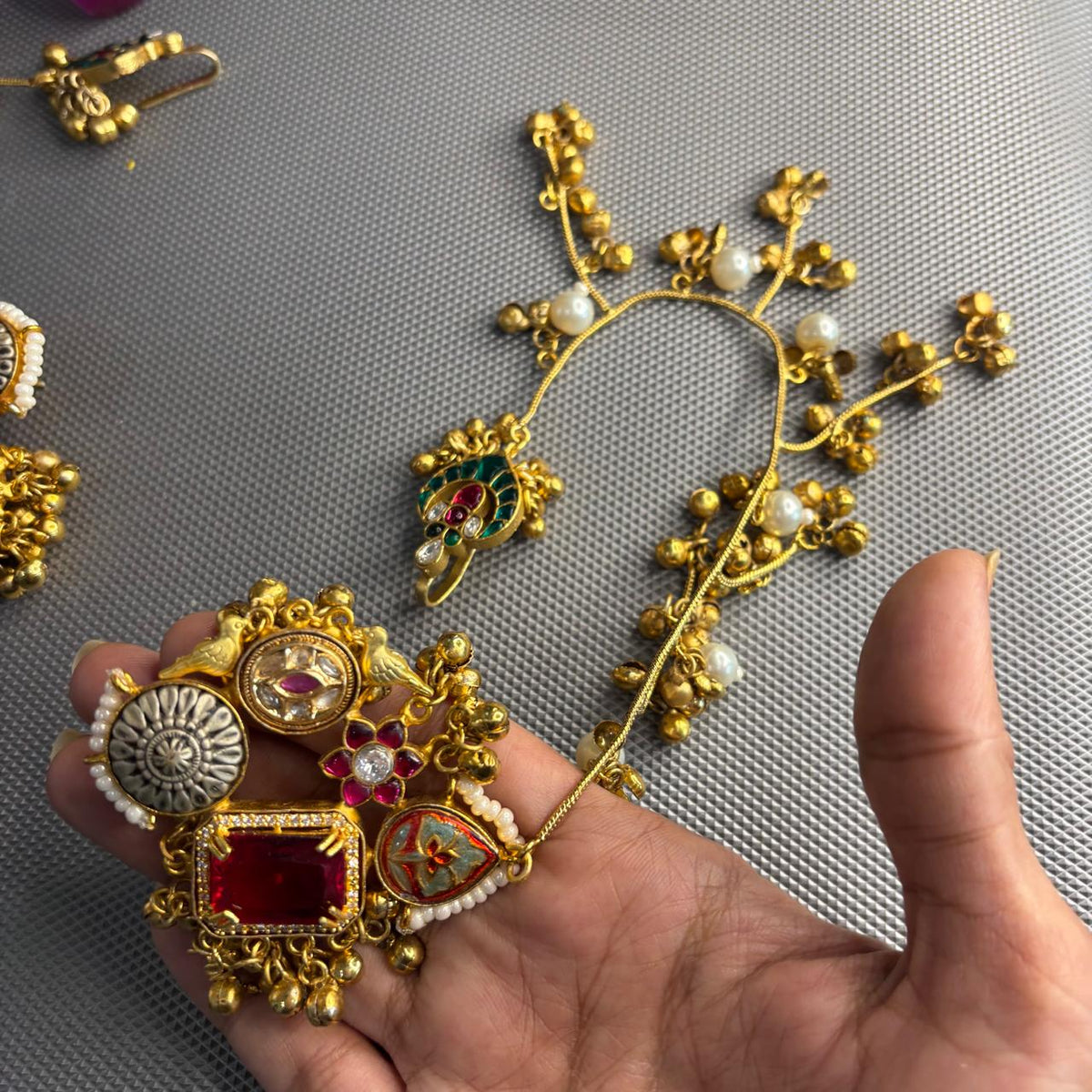 A close-up view of handheld gold-toned traditional  kashmiri Indian earrings with a matching earchain sahara  hair chain .The earring features a large red stone, a silver-toned circular motif, and small pearl-like beads.