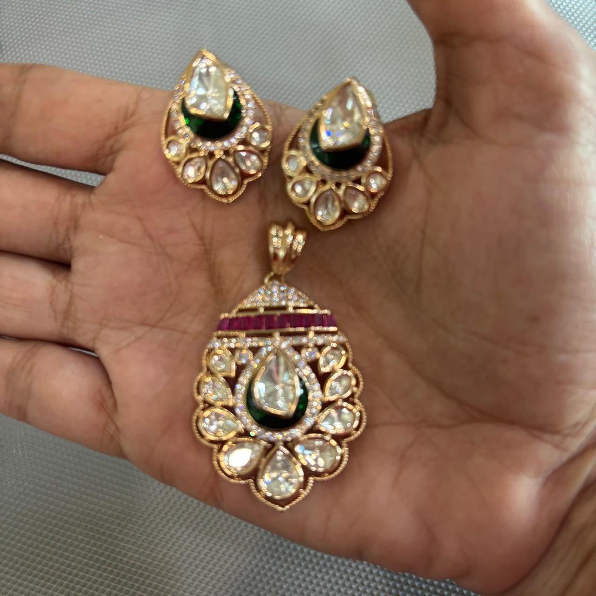 Gold-toned traditional Indian pendant  set featuring a teardrop-shaped pendant and matching stud earrings, embellished with clear stones, a dark green enamel , and a row of red stones, displayed on hand .