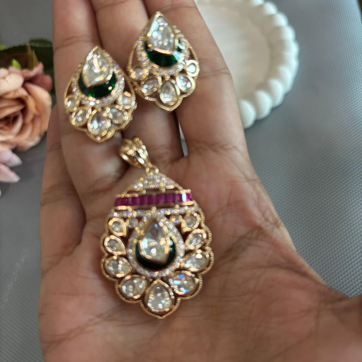 Closeup on hand a Gold-toned traditional Indian pendant  set featuring a teardrop-shaped pendant and matching stud earrings, embellished with clear stones, a dark green enamel , and a row of red stones . 