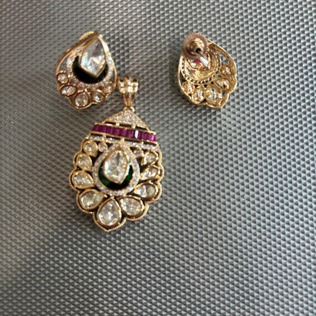 Gold-toned traditional Indian pendant  set featuring a teardrop-shaped pendant and matching stud earrings, embellished with clear stones, a dark green enamel , and a row of red stones , also showing the back side of the stud earrings.