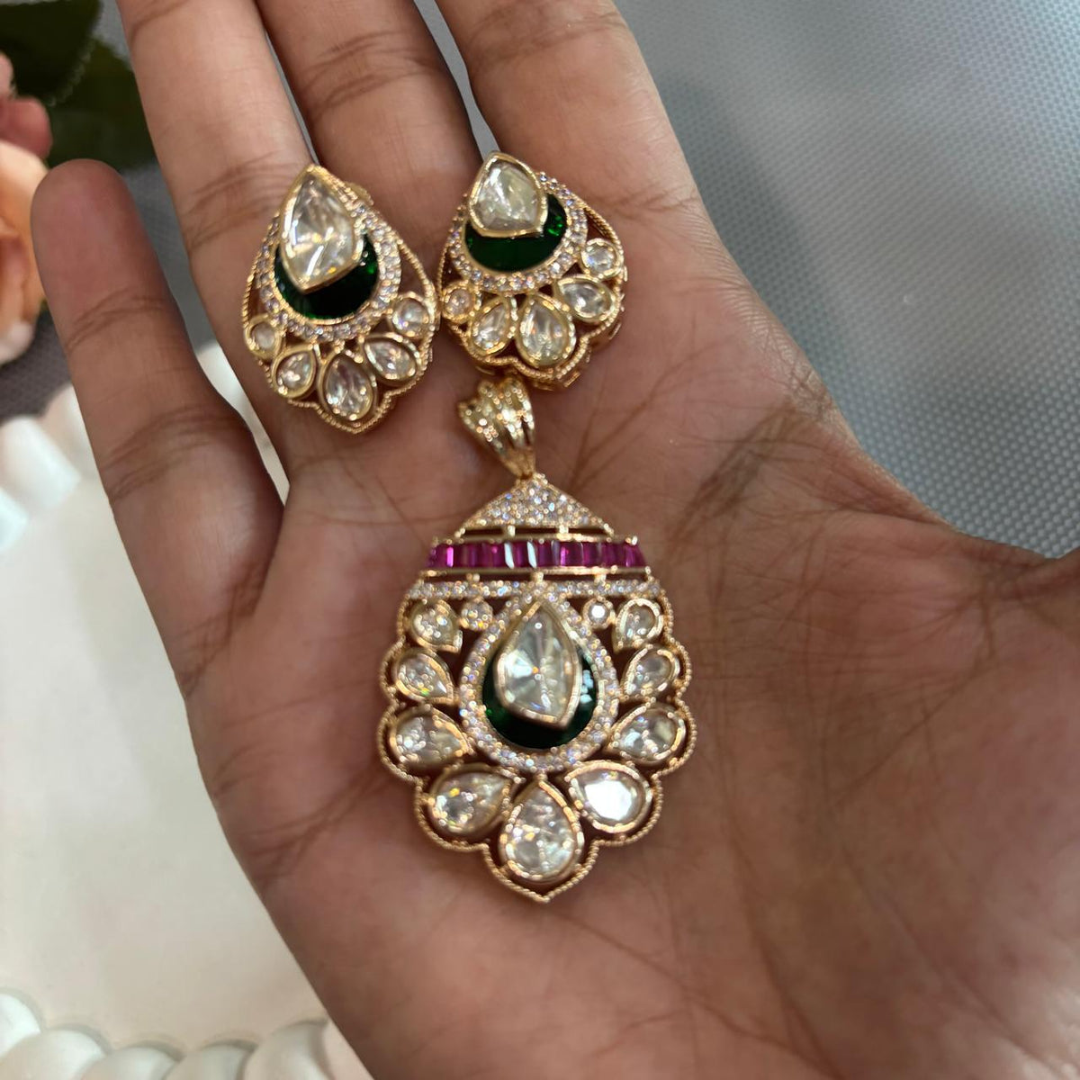 Hand held a Gold-toned traditional Indian pendant  set featuring a teardrop-shaped pendant and matching stud earrings, embellished with clear stones, a dark green enamel , and a row of red stones .